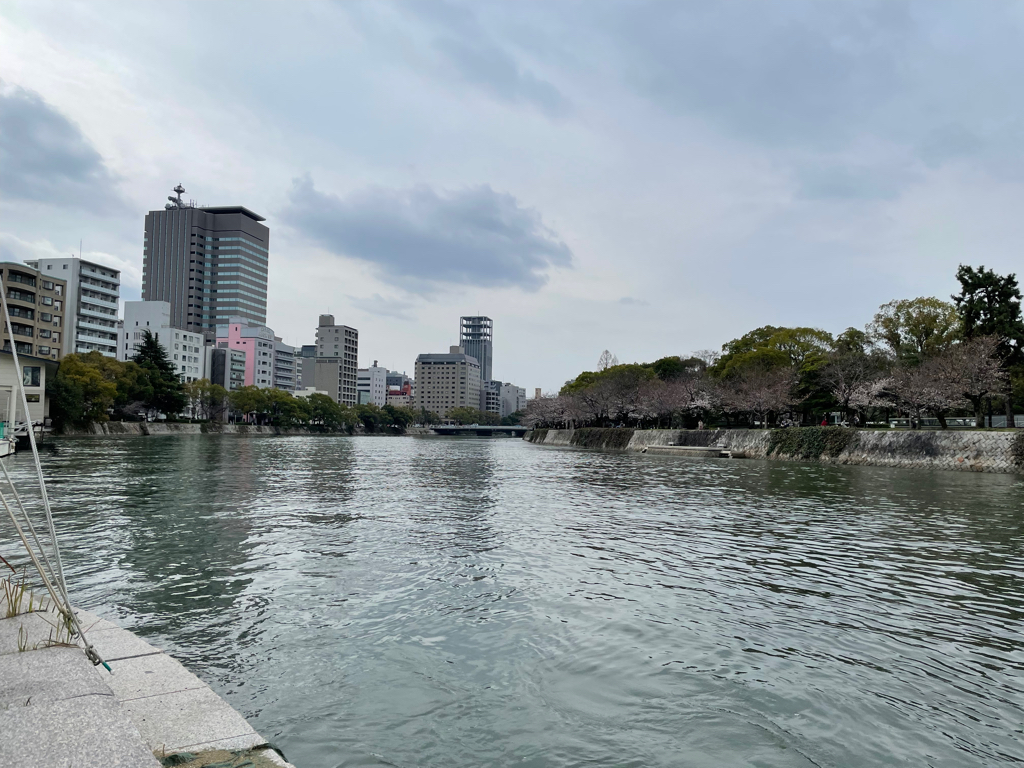 A view of a city waterfront with a river in the foreground. On the left side, there are several modern buildings, including some high-rise structures. On the right, there's a line of trees along the riverbank, some of which appear to be cherry blossoms. The sky is overcast with a few clouds.