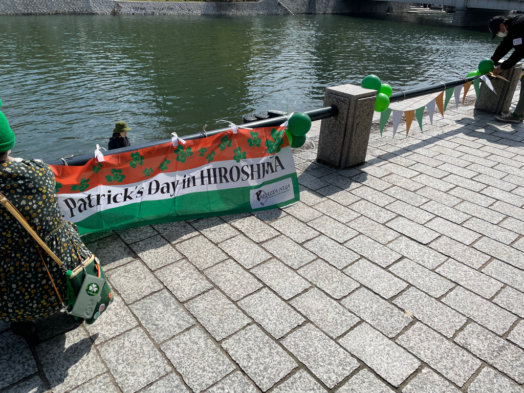 A banner hangs on a railing by a river, reading "Patrick's Day in Hiroshima" with clovers and green, white, and orange colors resembling the Irish flag. Green balloons are attached to the railing, and a person is fixing a string of triangular flags. Another person, with a green bag and wearing a floral-patterned outfit, is sitting nearby. The area is paved with stone tiles.