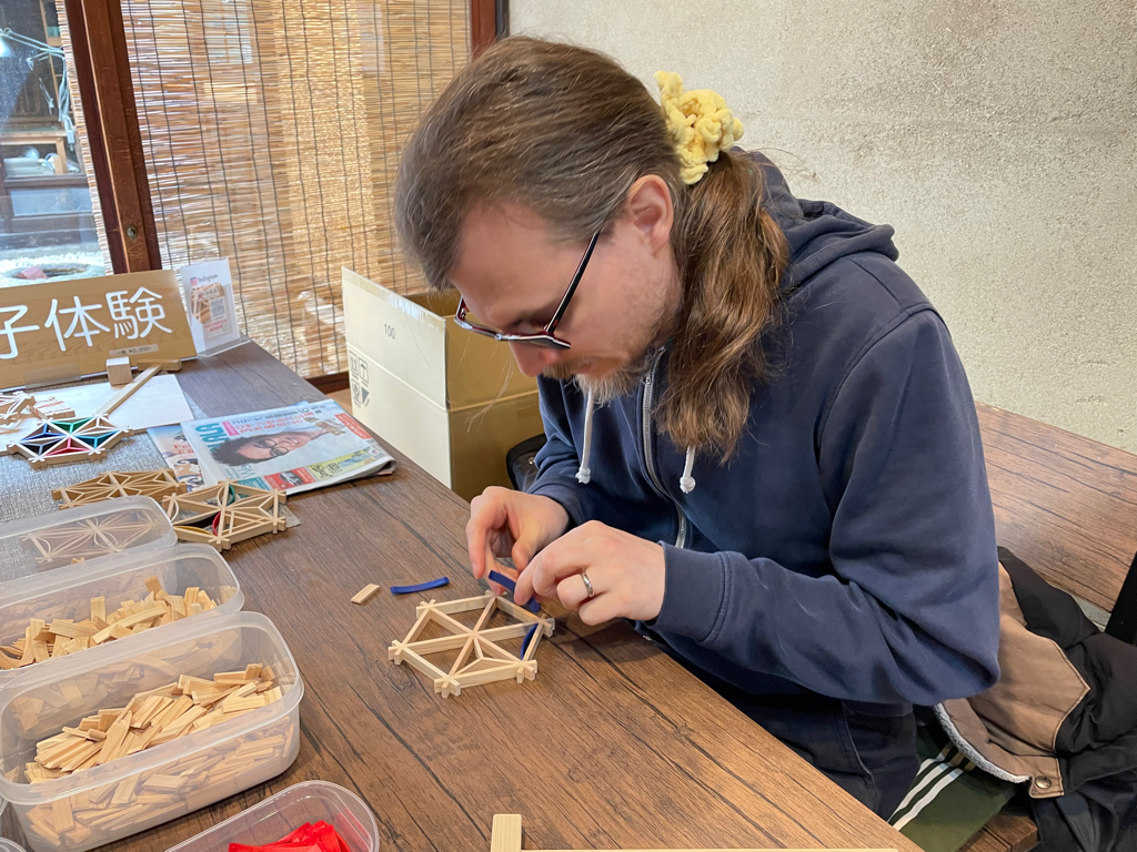 A person with long hair tied in a ponytail is concentrating on assembling a small wooden geometric structure at a table. The table has containers of wooden sticks and red fabric, and a partially assembled structure. A sign with Japanese characters is displayed nearby. The setting suggests a hands-on crafting activity.