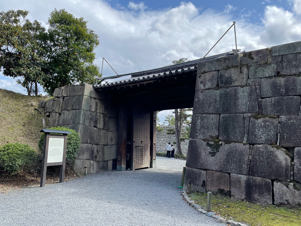 A large stone gateway with wooden doors stands open, leading into a historic site. The walls are constructed from large, weathered stones. There is a sign with information on the left side of the entrance (the content is not readable). Green bushes and a tree are visible near the gate, and the sky above is partly cloudy.