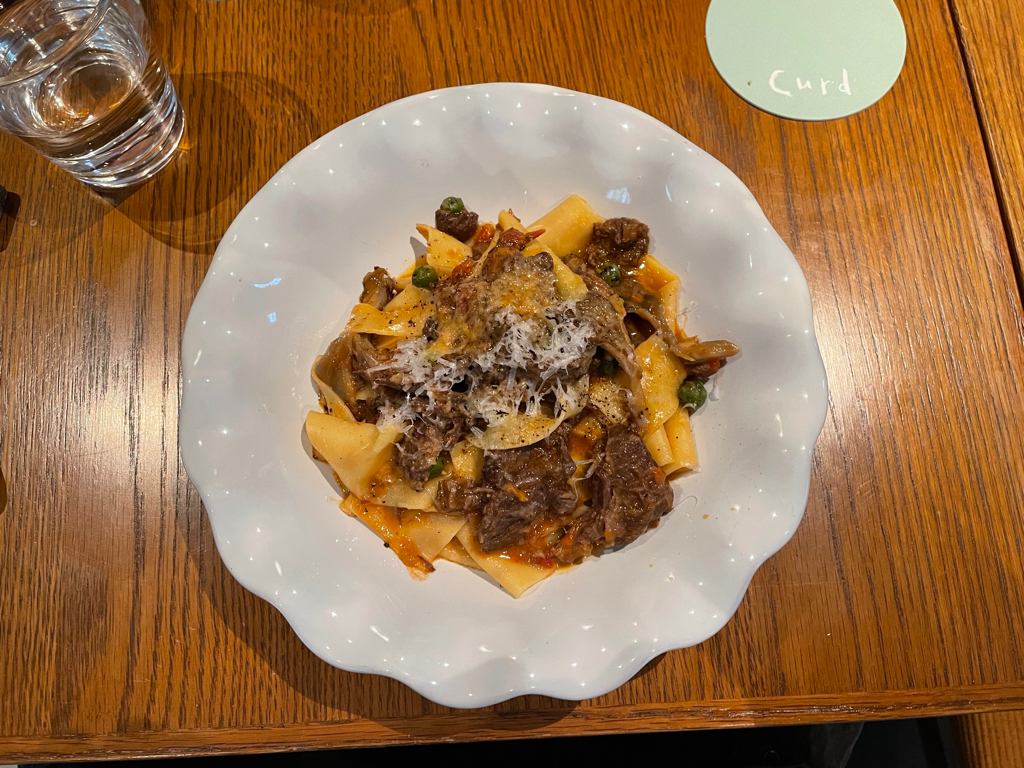 A plate of pasta topped with shredded cheese, pieces of beef, and vegetables is on a wooden table. The pasta is arranged on a white, scalloped-edge plate. Near the plate is a glass of water and a light green coaster with the text "Curd."
