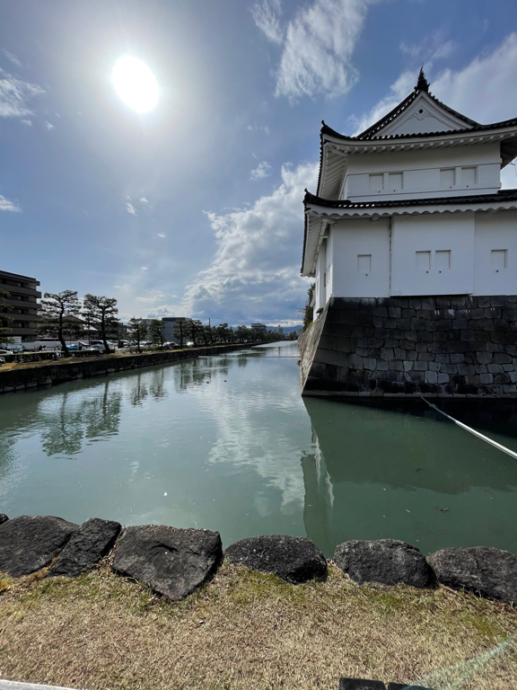A serene scene of a traditional Japanese building with white walls and a tiled roof, situated beside a calm body of water. The water reflects the structure and the sky, which is partly cloudy with the sun shining brightly. Trees and several buildings line the opposite bank of the waterway. A stone wall runs along the foreground, bordering a grassy area.