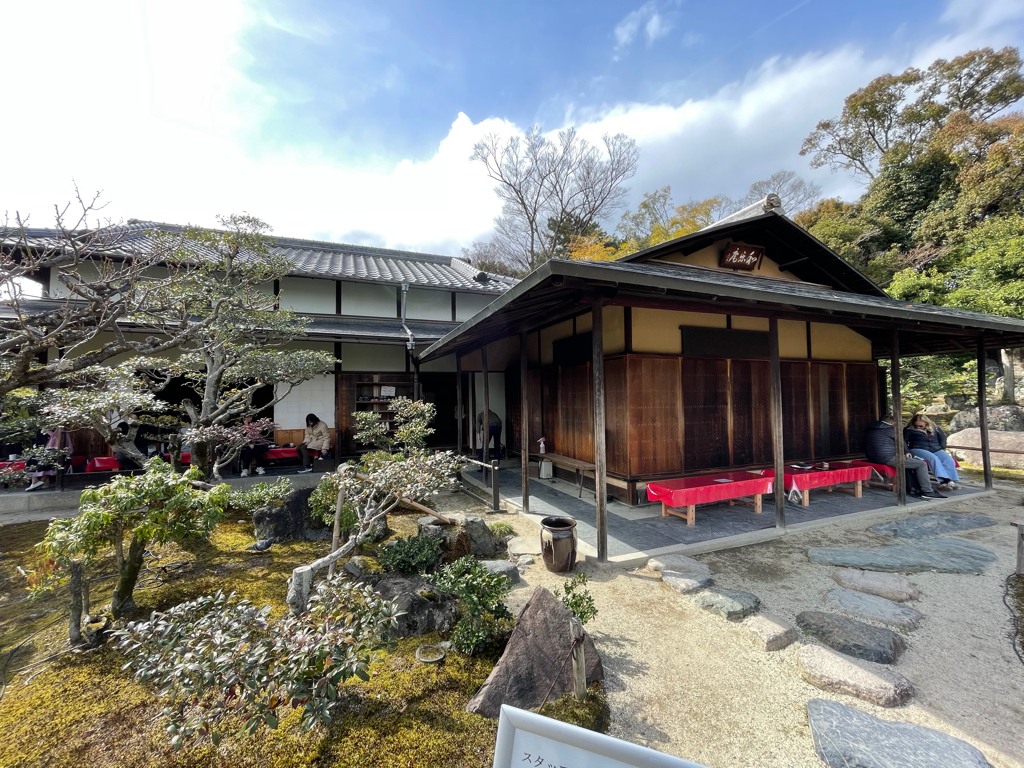 A traditional Japanese wooden building with a tiled roof, surrounded by a garden featuring carefully pruned trees and bushes. There are people sitting on benches with red cushions under the covered porch area.