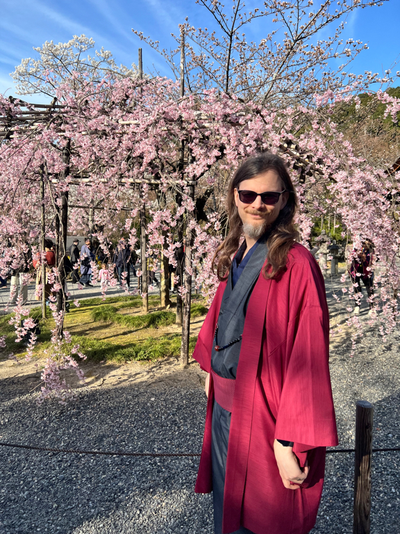 A person with long hair and sunglasses is wearing a traditional Japanese garment, featuring a red outer layer over a darker inner layer. They are standing in a garden with cherry blossom trees in full bloom, displaying pink flowers