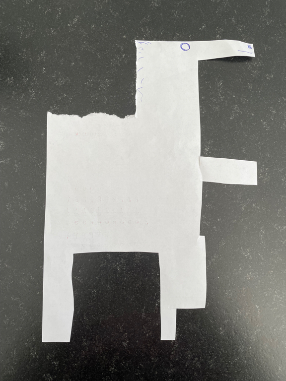 A piece of white paper has been cut into the shape of an abstract animal resembling a horse or a llama. It features a long neck, four legs, and a rectangular body. Simple facial details are drawn on it in blue pen, such as an eye and a few lines representing hair. The paper lays flat on a dark, textured surface.