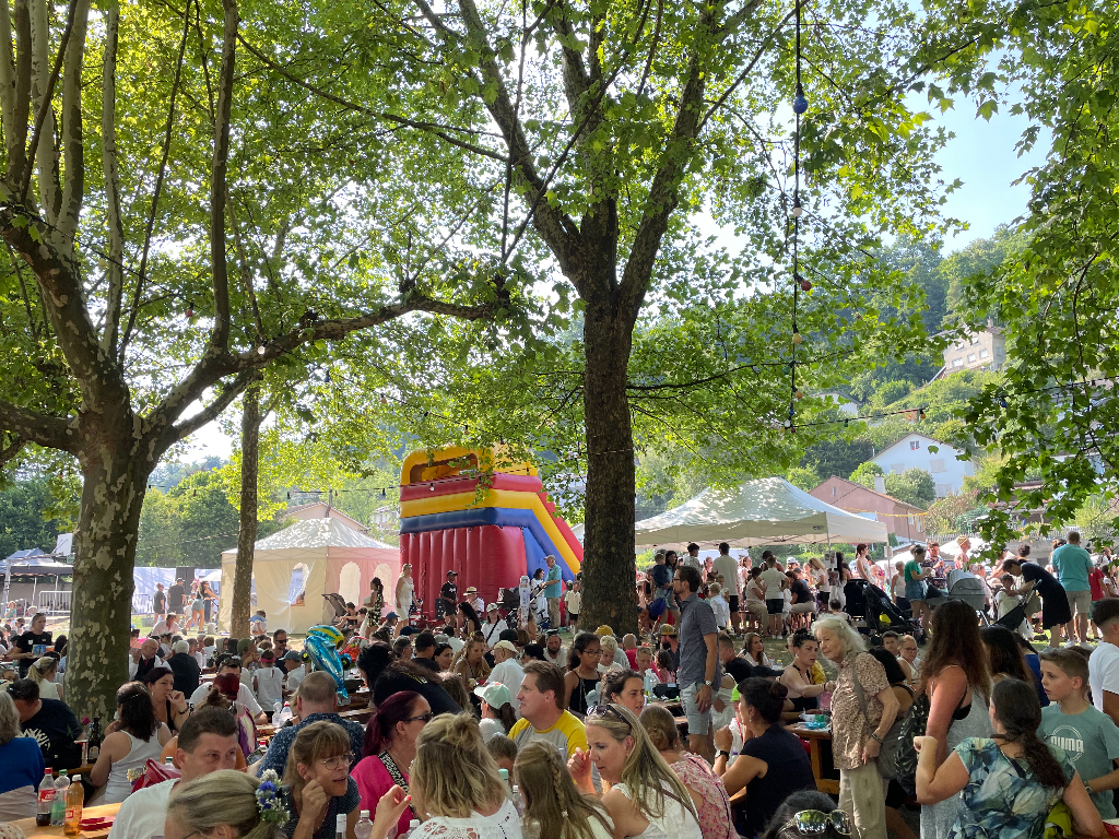 A lively outdoor festival scene under tall, leafy trees. Numerous people are gathered, seated at long tables, and enjoying various activities. A colorful inflatable play structure and several tents are visible in the background. The setting is bright and sunny, with greenery all around.