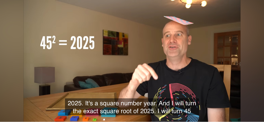 A man is sitting at a table inside a room, wearing a black T-shirt with a colorful design. There is a card balanced on his head. On the wall behind him, there's a painting. The text "45² = 2025" is displayed next to him, and the subtiles read: 2025. It's a square number year. And I will turn the exact square root of 2025 I Will turn, 45.
