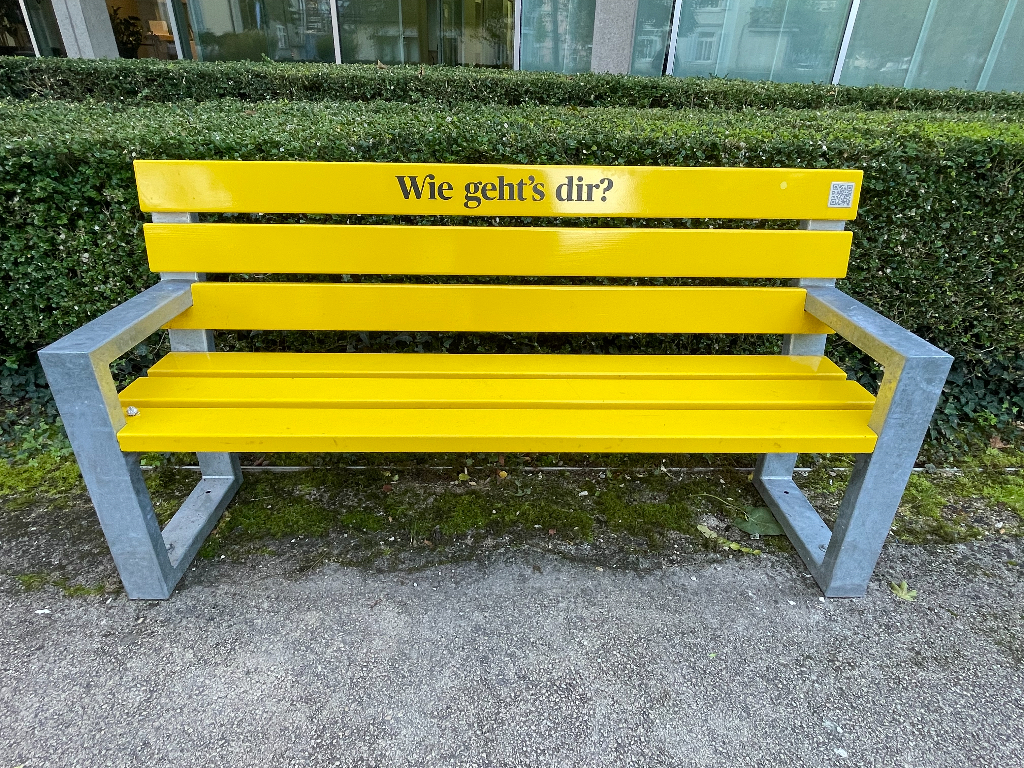 A bright yellow bench with gray metal frame is situated on a paved area surrounded by greenery. The backrest of the bench has the text "Wie geht’s dir?" printed on it, which is German for "How are you?". In the background, there are trimmed bushes and a building with large windows.