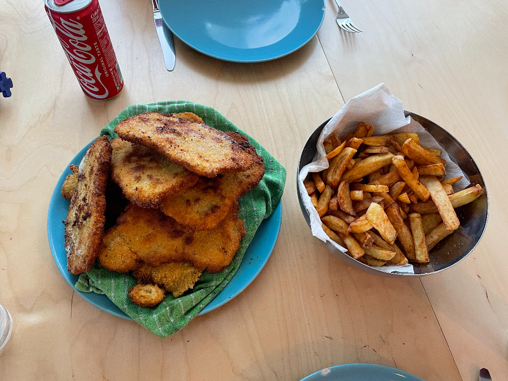 A plate of breaded and fried cutlets is on the left, placed on a green cloth on a blue plate. To the right is a metal bowl lined with paper, filled with golden-brown French fries. There is a can of Coca-Cola next to the plates. The setup is on a light wooden table with additional blue plates and cutlery visible in the background.