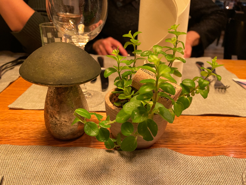 A table setting featuring a decorative stone or ceramic mushroom and a small potted plant with green leaves. The plant pot contains two corks. Behind them are a glass of water, utensils, and a folded white napkin. The table is covered with a gray placemat and there is a person in the background.