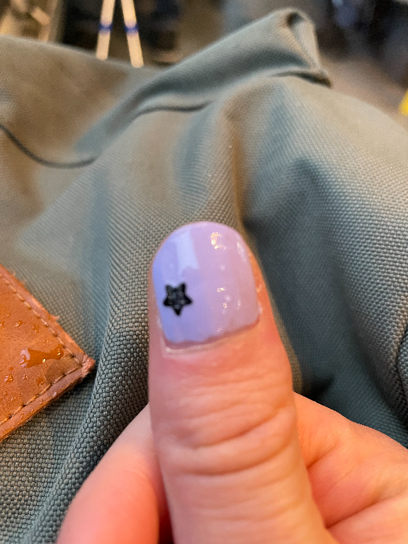 A close-up image of a thumb with a light purple, glossy nail polish. The nail is adorned with a small black star-shaped decoration near the cuticle. The background features a textured gray fabric and a small section of brown leather.