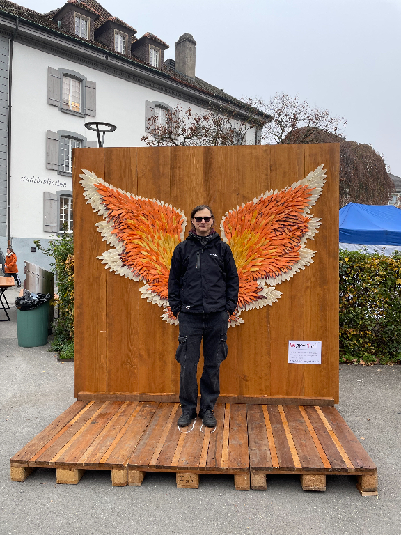 A person in dark clothing and sunglasses stands in front of a wooden backdrop with large, colorful wings painted on it, giving the appearance of having wings. The setting is outdoors on a cloudy day, with a building and some foliage in the background. The wings are orange, yellow, and white, designed to resemble feathers.