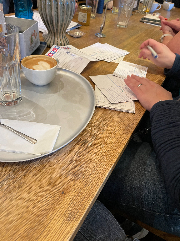 The image shows a wooden table with various items on it. There is a white tray holding an empty glass and a cup of coffee with a latte art heart in the foam, accompanied by a napkin and spoon. A person is visible on the right side, writing on several stacked pieces of paper with a pen. Additional items like a vase, glass, and jar are scattered in the background.