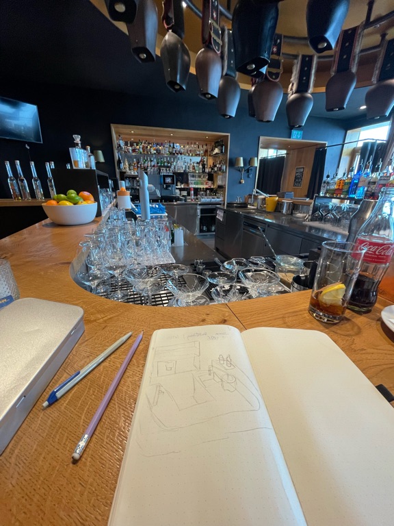A cozy bar interior with a curved counter featuring a variety of items. There's an open sketchbook with pencil drawings on the counter, beside some colored pencils. In the background, there's a shelf full of liquor bottles and glassware. A bowl of fruit, including oranges and limes, is placed on the counter. A glass with a lemon slice and a bottle of Coca-Cola are also present. Various ceiling-mounted beer taps hang above the bar. The lighting is warm and inviting.