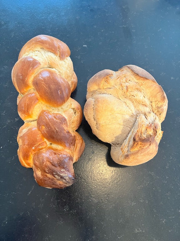 A close-up view of two loaves of bread on a dark surface. On the left is a golden brown braided loaf, and on the right is a round loaf with a rustic appearance.