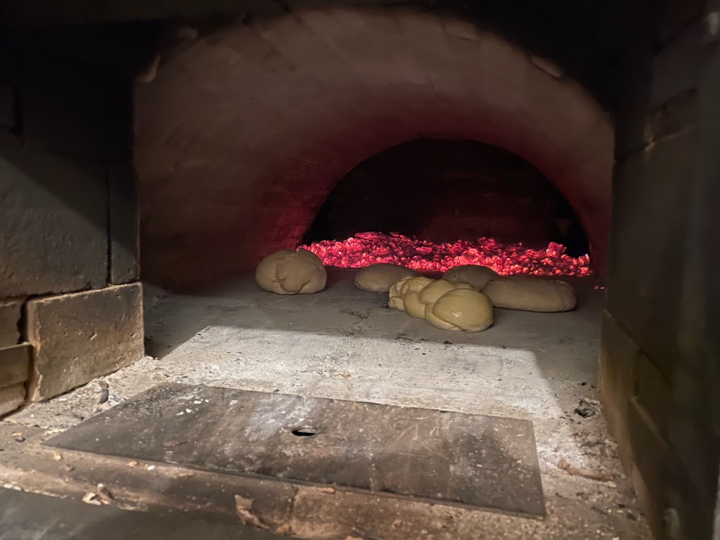 Dough balls are placed inside a large, traditional brick oven. The oven is heated with glowing red coals at the back, illuminating the dark interior with a warm glow.