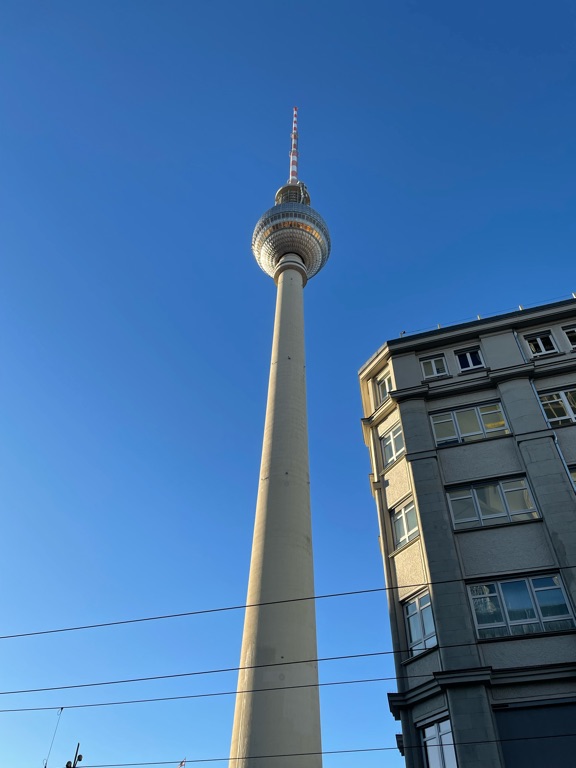 The image shows a tall TV tower with a spherical observation deck near the top, set against a clear blue sky. A section of a gray building with multiple windows is visible on the right side. The angle of the photograph captures the structures from below, emphasizing their height.