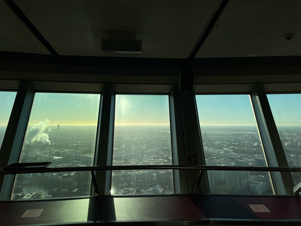View from inside a tall building through large windows overlooking a cityscape at sunset. The foreground shows a railing and part of the interior ceiling. A faint landscape of buildings is visible under a gradient sky transitioning from blue to orange. Smoke is rising from a chimney in the distance.