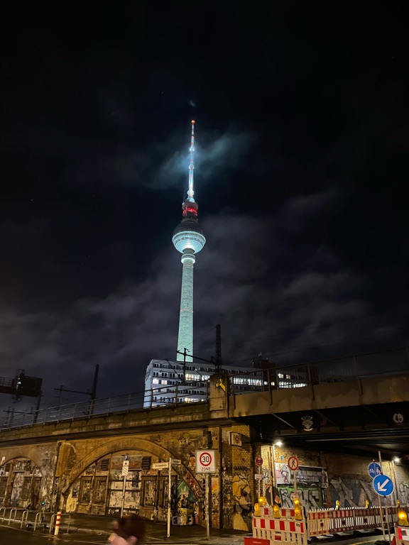 A nighttime scene featuring the Fernsehturm Berlin, a tall, illuminated tower with a glowing top, piercing through the clouds. Below, there's a graffiti-covered brick archway and urban structures, with a few street signs and barriers visible in the foreground. The sky is dark, enhancing the tower's bright presence.