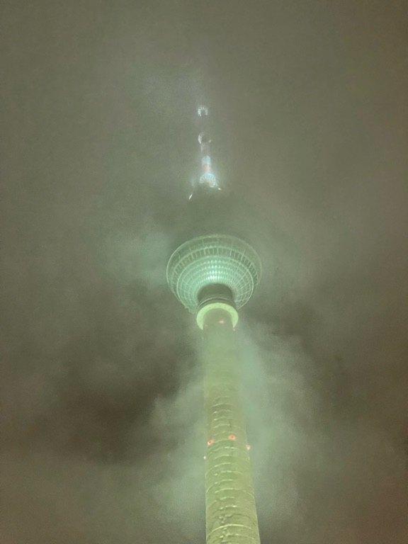 A tall telecommunications tower partially obscured by fog. The top of the tower is illuminated with greenish light, and the structure is shrouded in mist, creating a mysterious atmosphere.