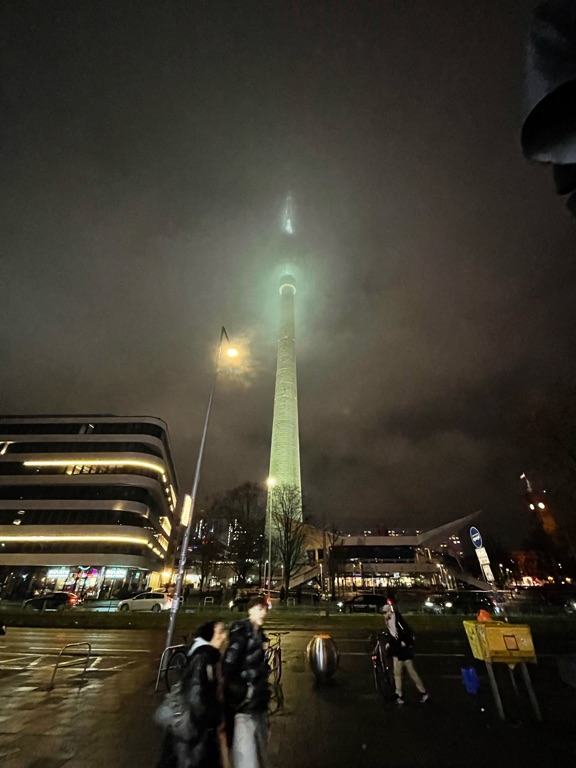 A nighttime city scene featuring a tall, illuminated TV tower with a glowing tip piercing through low clouds. The surrounding area includes a modern building with lit windows to the left and a street with people walking and biking in the foreground. The sky is overcast, adding a moody atmosphere to the image.