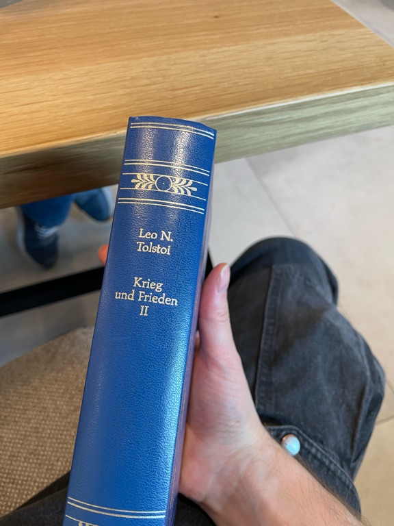 A person is holding a blue book with a spine that reads "Leo N. Tolstoi, Krieg und Frieden II" in gold lettering. The book is held against a light wood table. The person is wearing dark jeans, and their thumb is partially visible.
