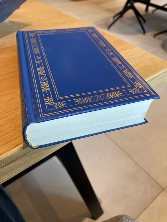A blue hardcover book with gold decorative patterns on the edges of its cover sits on a wooden table. The book is closed, and its pages are visible from the side. The table is in a room with a tiled floor and blurred furniture in the background.