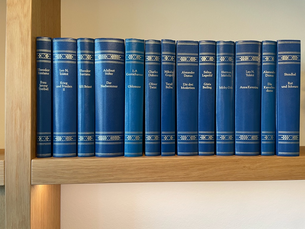 A row of classic, blue hardcover books with gold detailing on a wooden shelf. The titles, written in German, include "Frau Jenny Treibel," "Krieg und Frieden II," and "Die drei Musketiere." The spines are uniform in design.