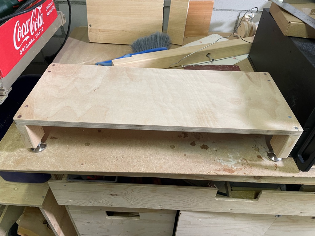 A wooden monitor riser with four legs is placed on a workbench. The riser is made of smooth, light-colored wood, supported by small metal brackets where the legs meet the surface. Behind it, there are various woodworking tools and materials, including a dust brush and some wooden planks. A red Coca-Cola box and other workshop items are visible in the background.