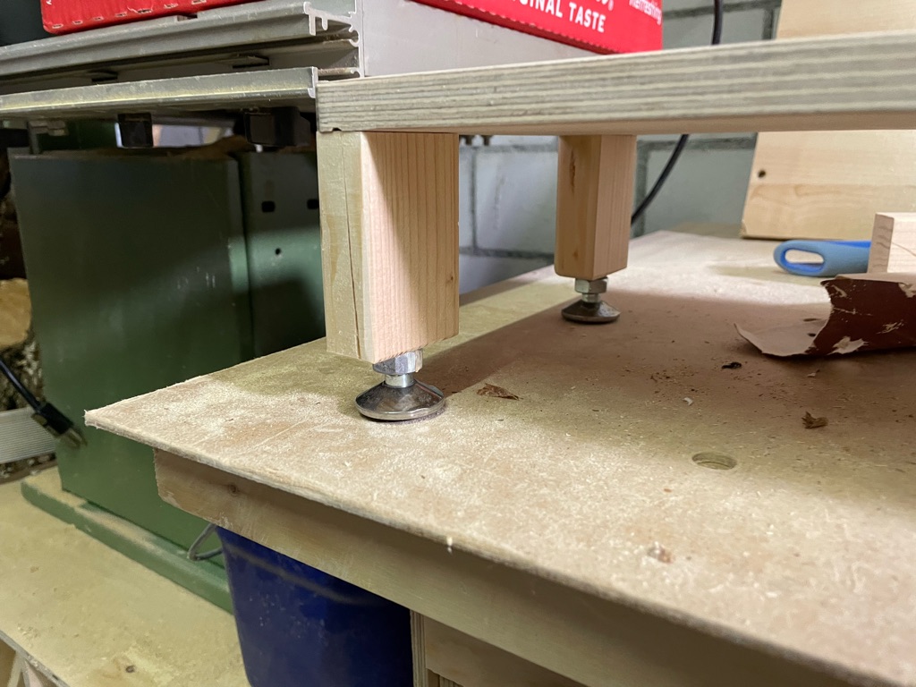 A wooden platform with adjustable leveling feet, made from threaded metal bolts and flat circular bases, is mounted on a workbench. The setup includes wooden legs attached underneath for support, and there's a piece of red packaging on the platform. The area is dusty, indicating woodworking activity, with tools and materials in the background.