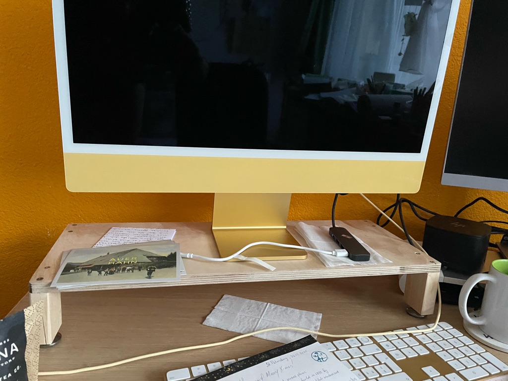 A workspace with a yellow desktop monitor on a wooden stand. Below the monitor are note papers and a device with cables attached. To the right, part of another screen and some electronic devices are visible. A white keyboard is partially shown at the bottom, and a white mug is situated on the desk. The wall in the background is painted orange.