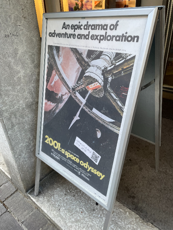 A poster displayed on an A-frame stand outside a building features an image from the movie "2001: A Space Odyssey." The text at the top reads "An epic drama of adventure and exploration," with the movie's title in bold yellow letters at the bottom. The poster includes an image of a spacecraft and a planet, with a QR code visible.