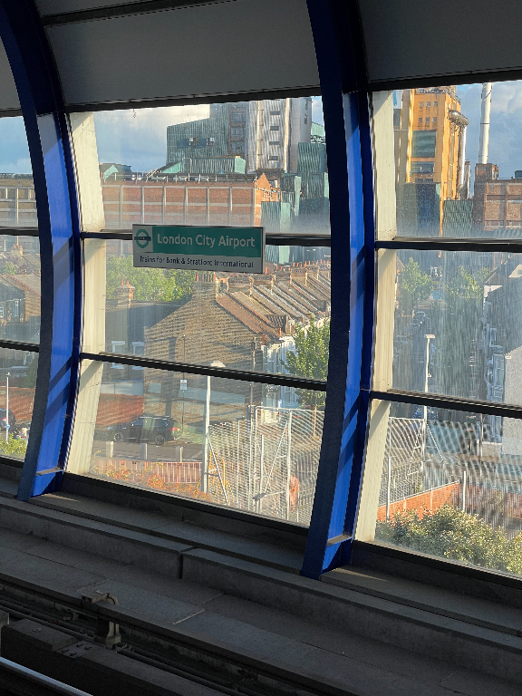 View from a train platform at London City Airport station, looking out through large curved windows. The backdrop includes urban buildings with a mix of modern and older architecture. A sign indicating the station name is visible against the cityscape, and the platform is empty with no people present.