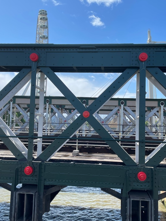 View of a bridge with metal beams in a crisscross pattern, highlighted by red bolts. The ferris wheel is visible in the background against a clear blue sky, and water flows under the bridge.