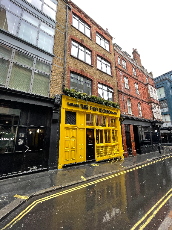 A city street features buildings with varied architectural styles. The central building has a striking yellow facade with large windows and a sign above the door. It contrasts with the adjacent buildings, one with a modern design and another with classic brickwork. The road is wet, reflecting the buildings, indicating recent rain.