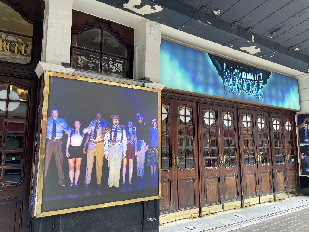 The image shows the exterior of a theater featuring a large poster of a theatrical production. The poster depicts a group of actors in costume on stage, standing in a line under colorful lighting. Above the theater's wooden doors, there is a marquee with the title of the production in stylized lettering against a blue and green background. The theater's entrance is ornate, with glass-paneled wooden doors.