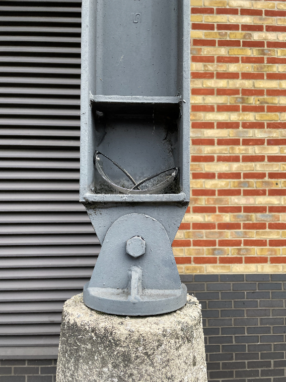 A close-up of a metal structure, mounted on top of a concrete base. Inside the metal beam are a safety glasses. The structure has a bolt and sits in front of a brick wall. The background are louvered metal strips on the left and yellow brick on the right.