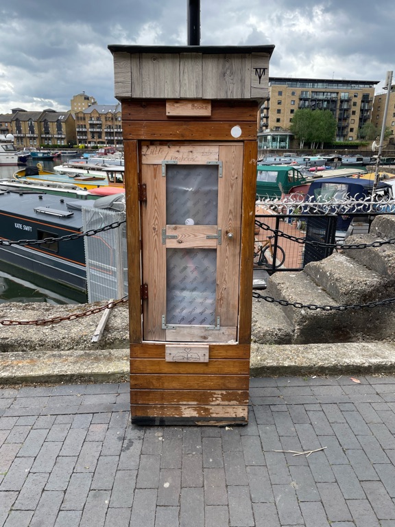 A small wooden structure resembling a phone booth is situated on a paved walkway by a waterfront. The booth has a clear window with a wooden frame and appears weathered. In the background, there are moored boats of various colors and buildings along the water's edge. The sky is cloudy, casting a subdued light over the scene.