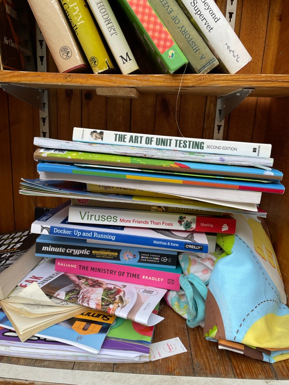 A wooden bookshelf filled with assorted books. The most prominent title visible is "The Art of Unit Testing Second Edition." Other book titles include "Viruses: More Friends Than Foes," "Docker: Up & Running," and "Minute Cryptic." A few colorful children's books and magazines are also present. Some books are stacked horizontally, and a soft, colorful fabric is draped on the side.