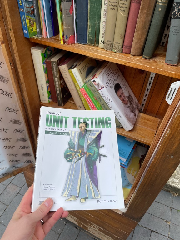 A person is holding a book titled "The Art of Unit Testing" by Roy Osherove in front of a wooden bookshelf filled with various books. The cover features an illustration of a medieval character in colorful robes holding a quill and scroll. Some books are stacked vertically and horizontally on the shelves. The ground is paved with gray stone tiles.