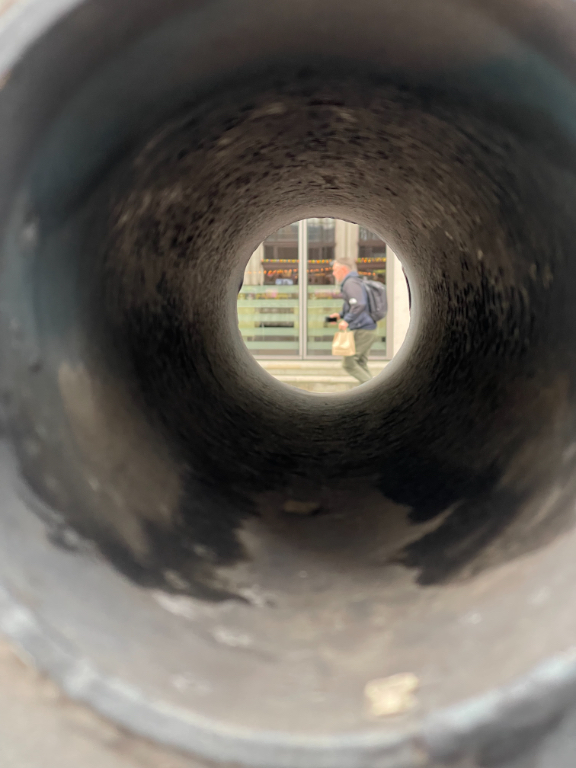 View through a black pipe with a blurred figure of a person walking on the sidewalk outside. The scene looks circular from the perspective of the pipe's interior, with the exterior appearing in the background.