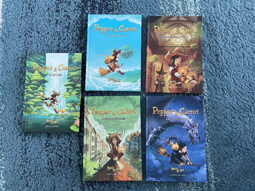 A collection of five "Pepper & Carrot" comic books laid out on a textured blue surface. Each book features colorful cover art showcasing a whimsical fantasy theme, with illustrations of a young witch on a broomstick, her orange cat, magical creatures, and various fantastical settings. The titles and illustrations suggest an adventurous and magical storyline.