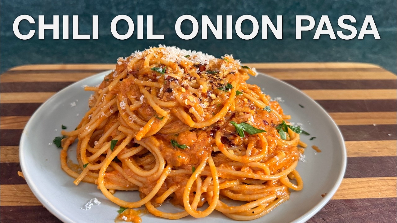 Alt text: A YouTube video thumbnail featuring a plate of spaghetti topped with a rich, orange-red sauce, garnished with chopped herbs and grated cheese. The text “CHILI OIL ONION PASTA” is displayed at the top of the image. The dish is placed on a wooden surface.