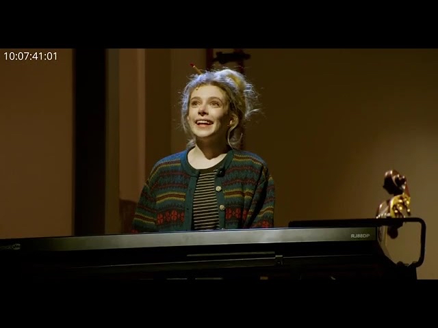 A person with light hair is standing and playing a keyboard in a dimly lit room. They are wearing a colorful sweater over a striped shirt, with their hair in a bun. The timecode “10:07:41:01” is visible in the upper left corner.