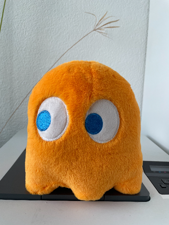 A plush toy resembling an orange ghost with large white eyes and blue pupils, sitting on a black device. The background features a light-colored wall and a small part of a plant.