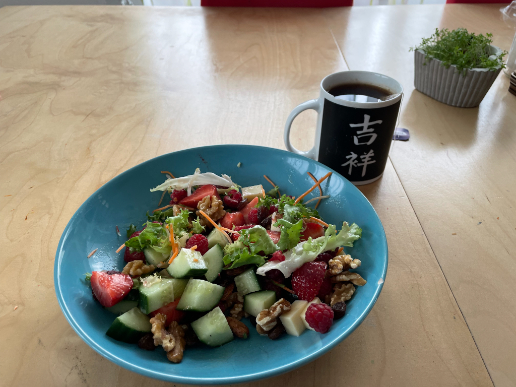 A bowl of salad on a wooden table, containing lettuce, cucumbers, strawberries, raspberries, walnuts, and shredded carrots. Next to the bowl is a mug with Asian characters on it, filled with a dark beverage. There is also a small potted plant in the background.