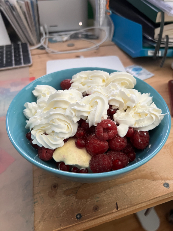 A bowl of raspberries topped with swirls of whipped cream, placed on a wooden table. In the background, there are office supplies, including a keyboard, folders, and stacks of paper.