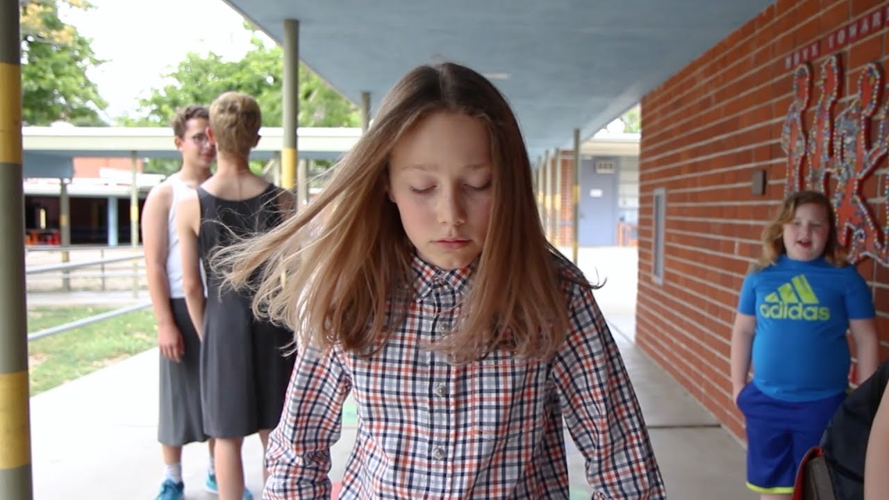 A YouTube video thumbnail shows a young person with long hair and a plaid shirt walking with a downcast look in a school corridor. In the background, there are two other people standing close together having a conversation, and another person wearing a blue shirt with an &ldquo;Adidas&rdquo; logo is walking towards them. The scene takes place outdoors under a covered walkway next to a brick wall.