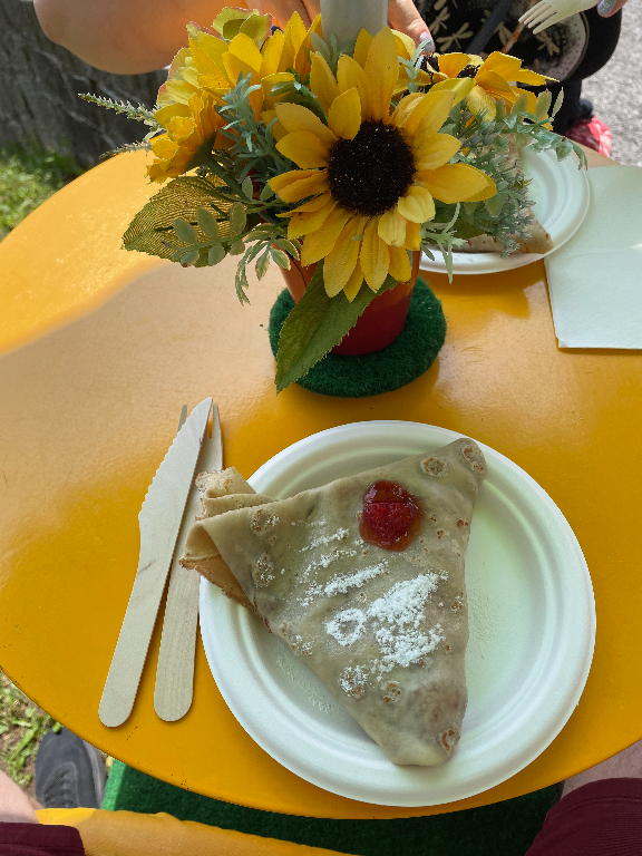 A folded crepe with powdered sugar and a dollop of jam on a white plate is placed on a bright yellow table. Next to the plate are wooden utensils. In the background, there is a small pot of sunflowers in a red container, adding a cheerful touch to the setting.