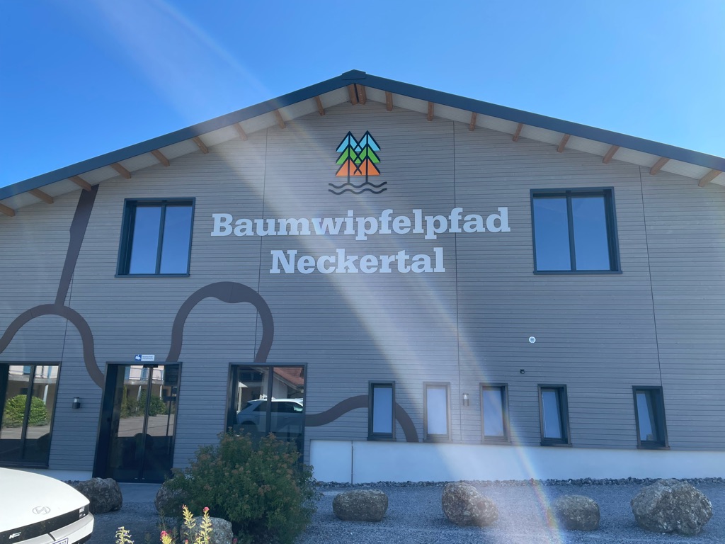 The image shows the exterior of a building with the words "Baumwipfelpfad Neckertal" on the side. The building has a wooden facade with two large windows and several smaller ones. There is a colorful logo featuring three stylized trees above the text. The sky is clear and blue, and some shrubbery is visible near the front entrance. The sunlight creates a diagonal lens flare across the image.